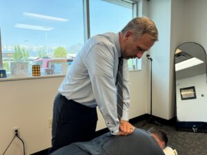Chiropractor in light blue shirt and tie adjusting patient's lower back in modern office with mountain view